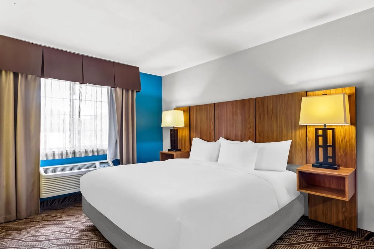 Quarto de hotel com uma cama grande, duas luminárias de cabeceira, uma janela com cortinas e um aquecedor fixado na parede.