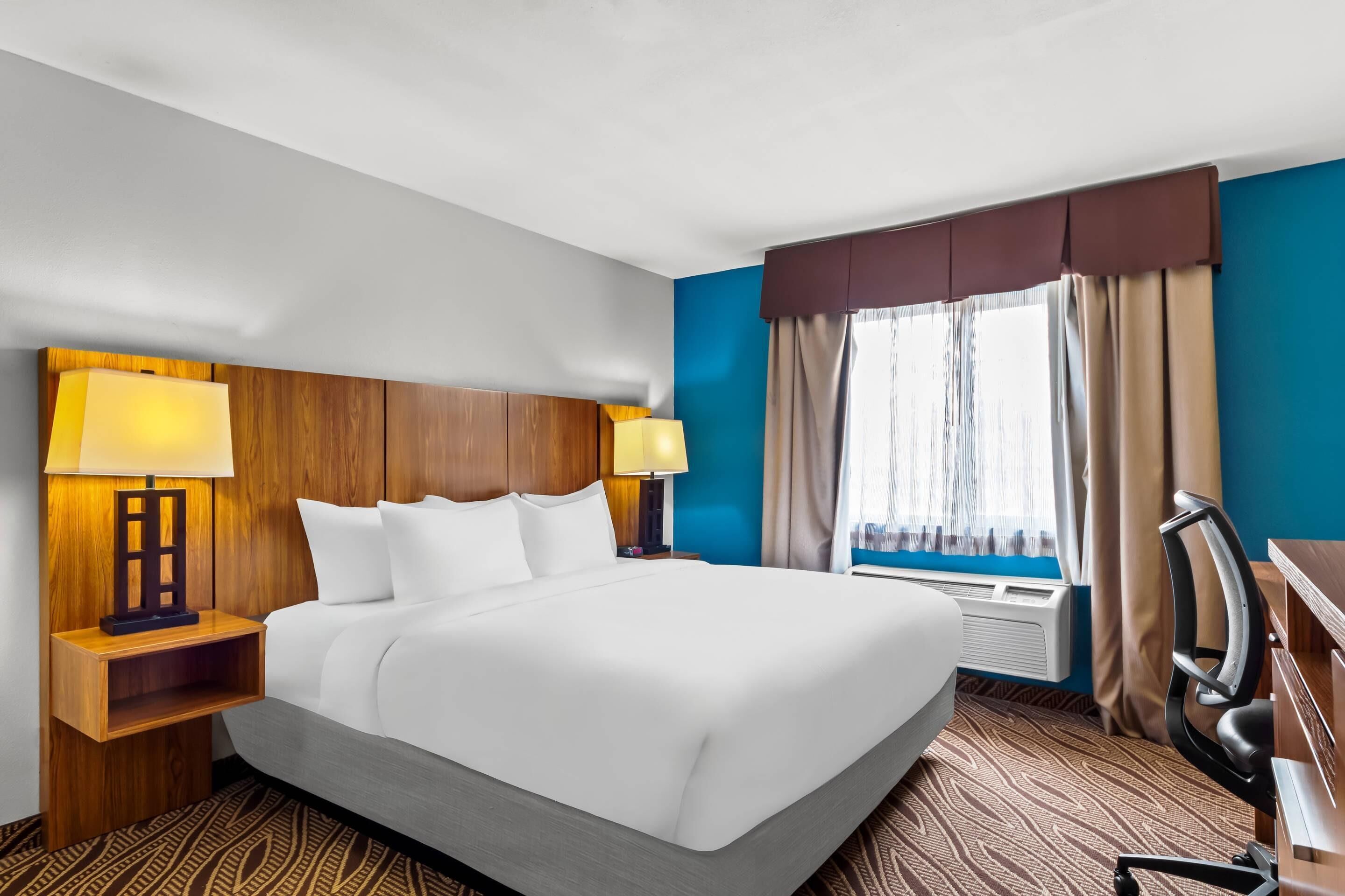 A hotel room with a large bed, two bedside tables with lamps, a desk with a chair, and a window with curtains.