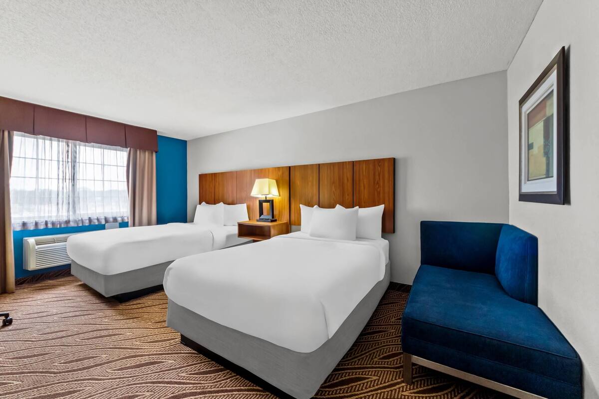 Une chambre d’hôtel avec deux lits, un fauteuil bleu, une tête de lit en bois et une fenêtre avec des rideaux.