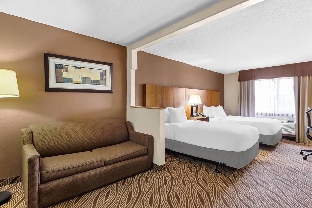 Une chambre d’hôtel avec un canapé marron, deux lits, un tableau encadré au mur et une moquette à motifs.