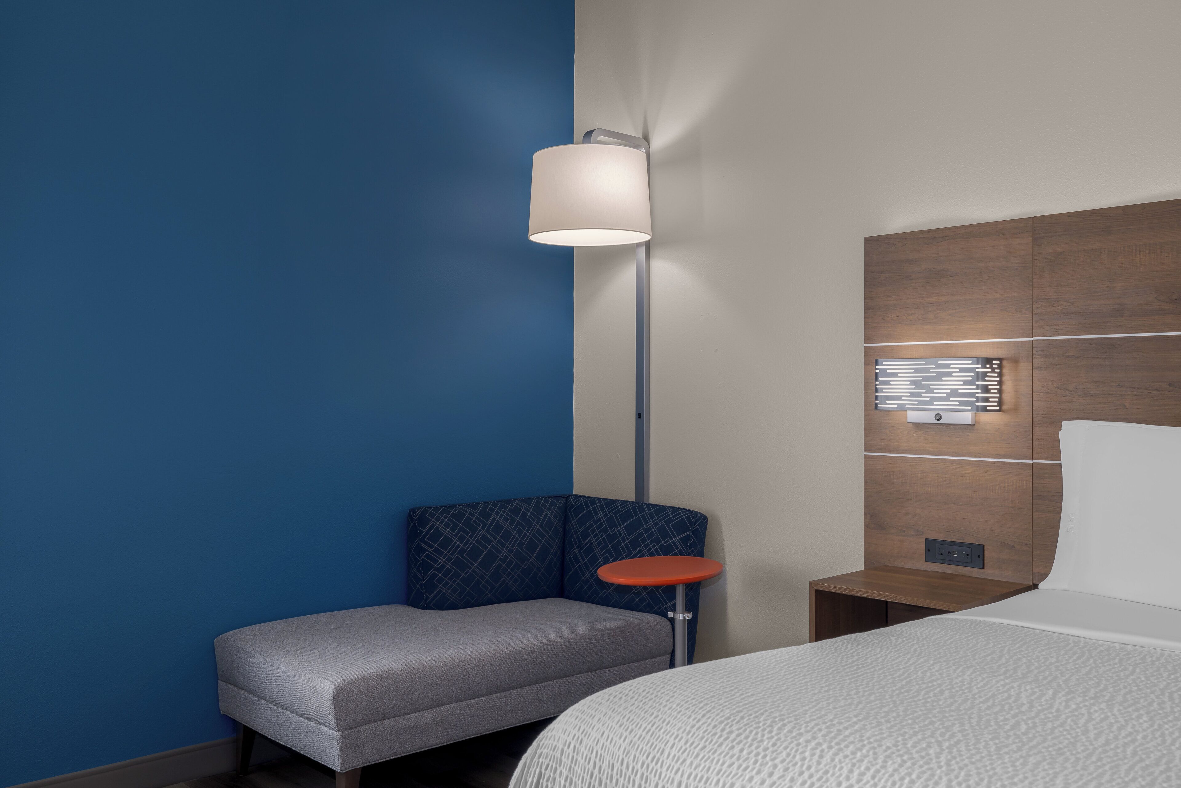 Habitación de hotel con cama, mesita de noche, lámpara, una pequeña mesa y una pared azul decorativa.
