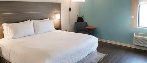 Un lit bien fait, avec du linge de lit blanc et un motif géométrique, une table de chevet avec une lampe, une chaise et une petite table avec une lampe.