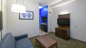 Una habitación de hotel moderna con sofá, una mesa de madera, un televisor sobre un mueble y una zona de cocina al fondo.
