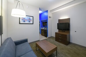 Une chambre d’hôtel moderne avec un canapé, une table en bois, un téléviseur posé sur un meuble et un coin cuisine à l’arrière-plan.