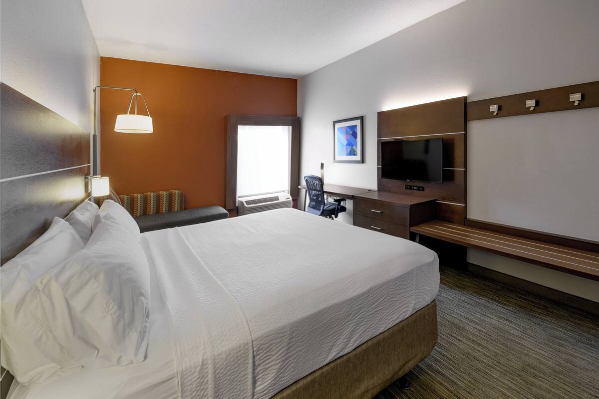 A hotel room with a large bed, a desk with a chair, a flat-screen TV, and a window with blinds.