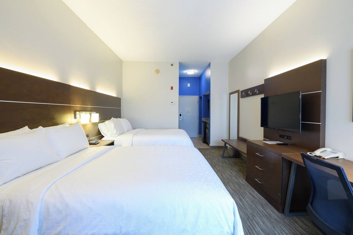 A hotel room with two beds, a desk with a television, and a small kitchenette.