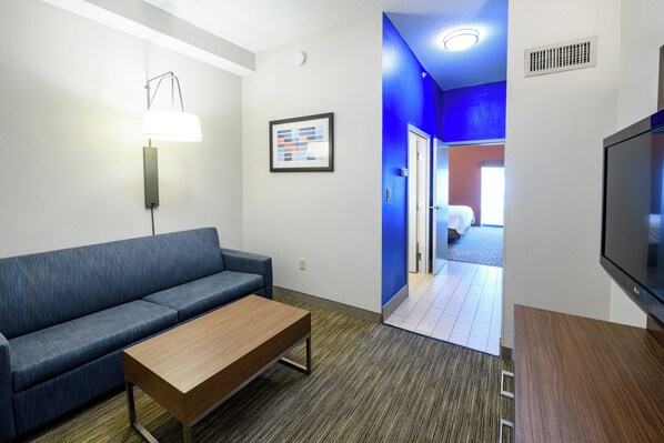 Une chambre d’hôtel moderne avec un canapé bleu, une table basse en bois, un téléviseur à écran plat et un tableau encadré au mur.