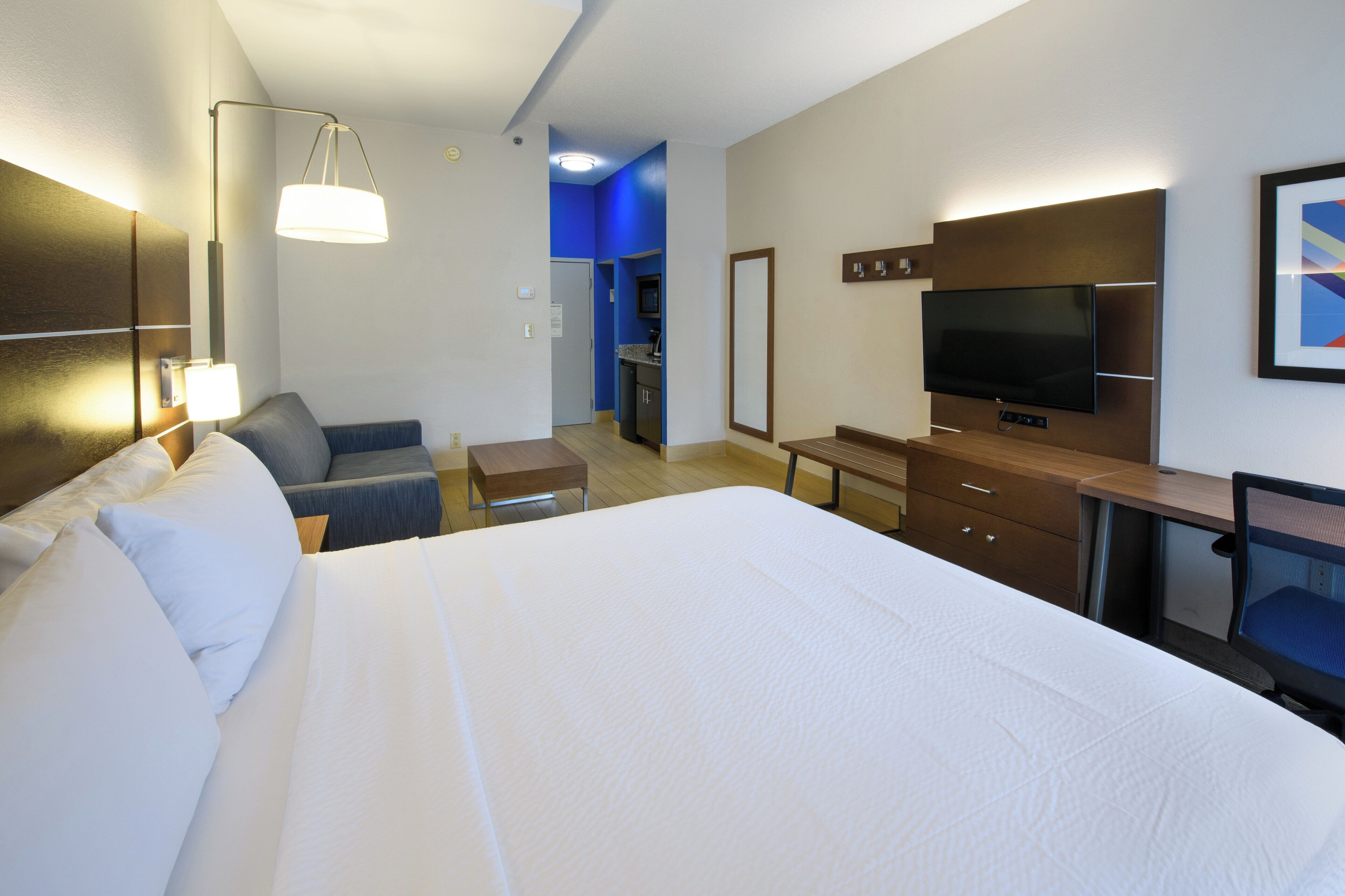 Une chambre d’hôtel avec un grand lit, un bureau, une chaise, une télévision et, au fond, une petite cuisine.
