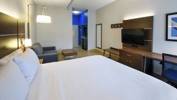 Habitación de hotel con una cama grande, un escritorio, una silla, un televisor y una kitchenette al fondo.