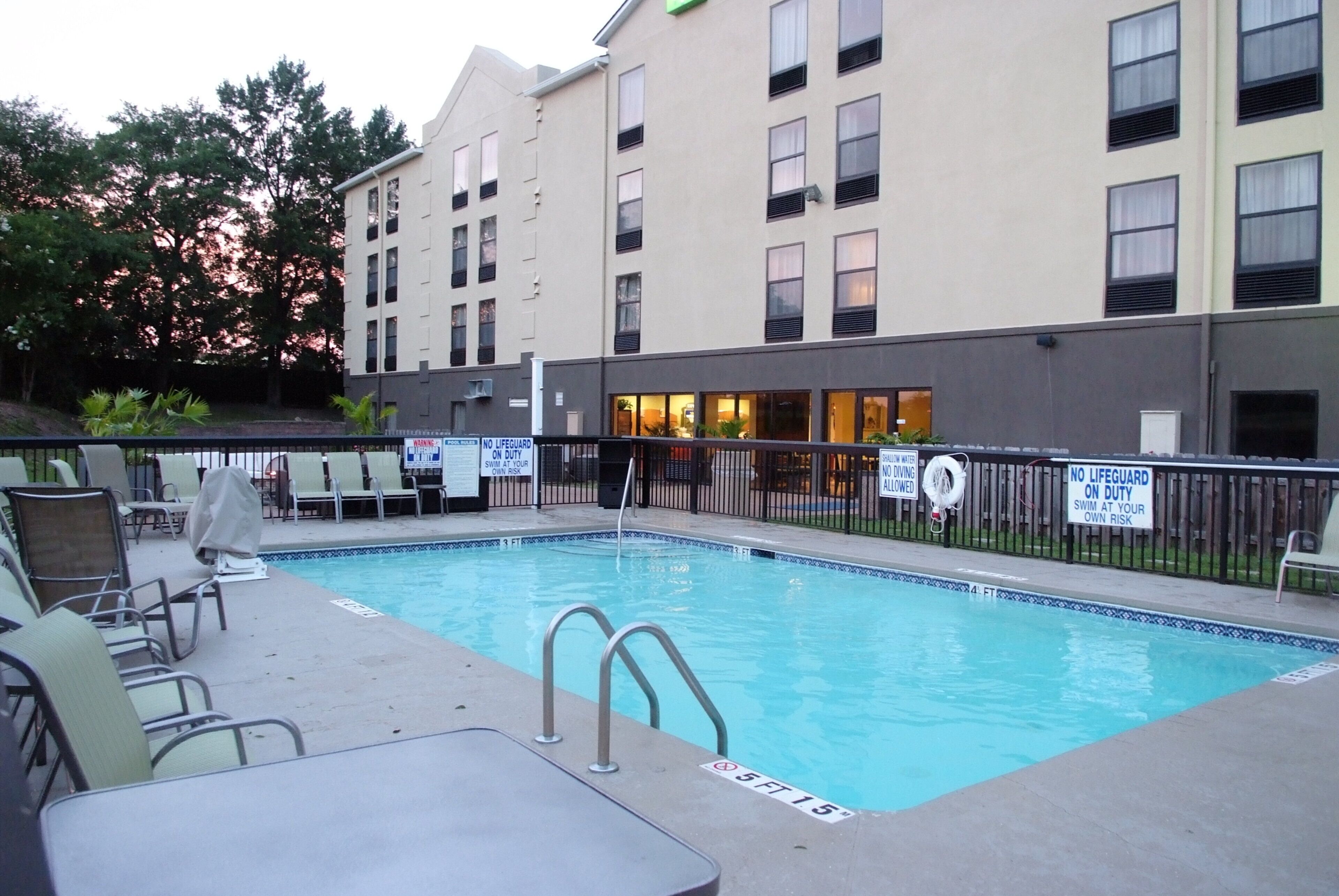 A swimming pool with no lifeguard on duty, surrounded by lounge chairs and a building with multiple windows.
