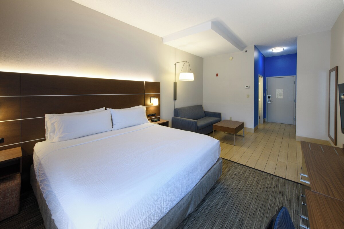 Une chambre d’hôtel avec un grand lit, des tables de chevet, un bureau, une chaise et une salle de bain visible à travers une porte ouverte.
