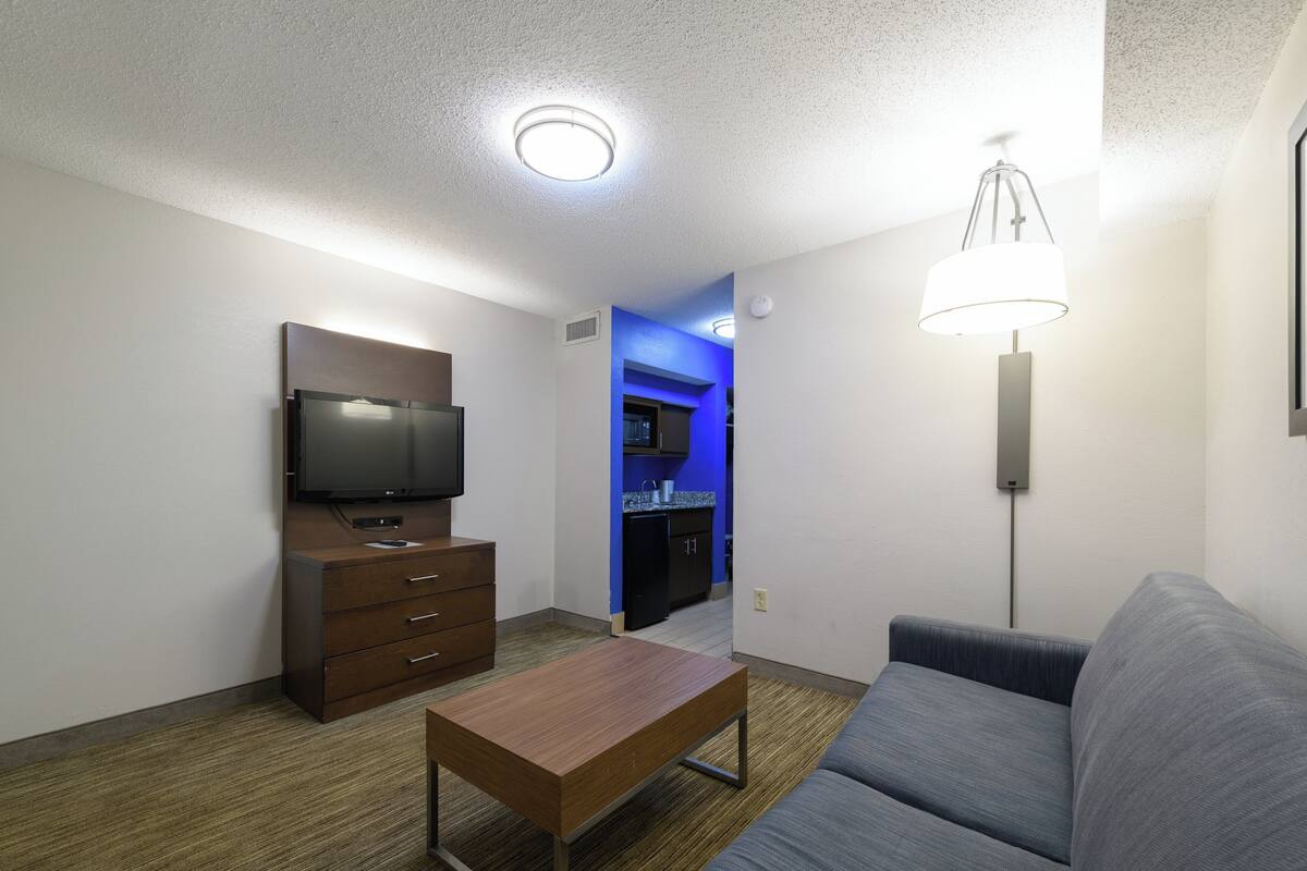 A modern hotel room with a flat-screen TV, a wooden coffee table, a gray sofa, and a standing lamp.