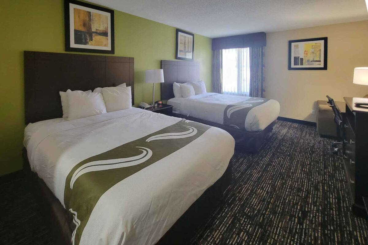 A hotel room with two beds, a desk, a chair, and a lamp.