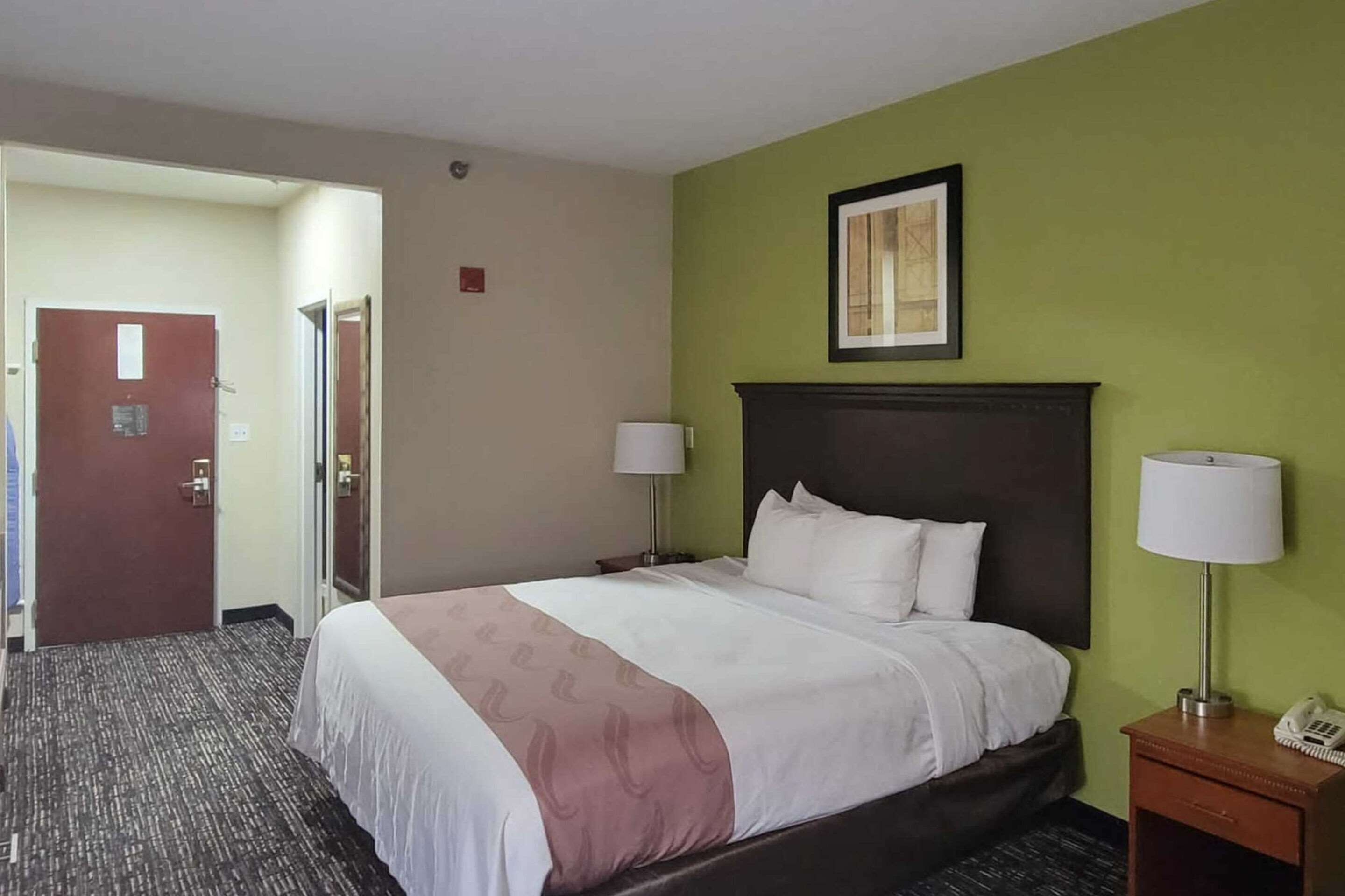 Una camera d'hotel con un letto, due lampade da comodino, un comodino e una porta che dà su un'altra stanza.