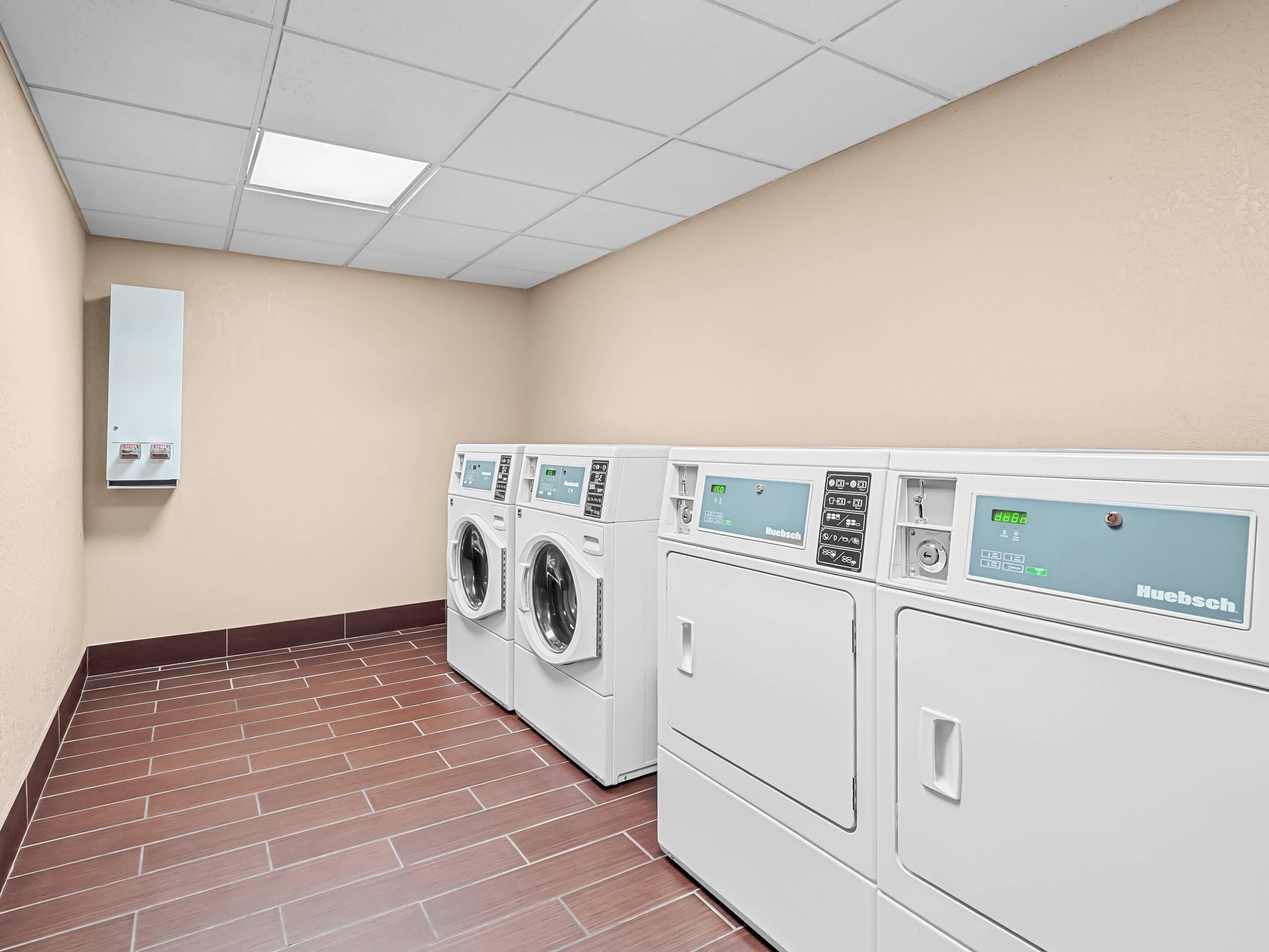 A laundry room with four white Huebsch washing machines, a beige wall, and a tiled floor.