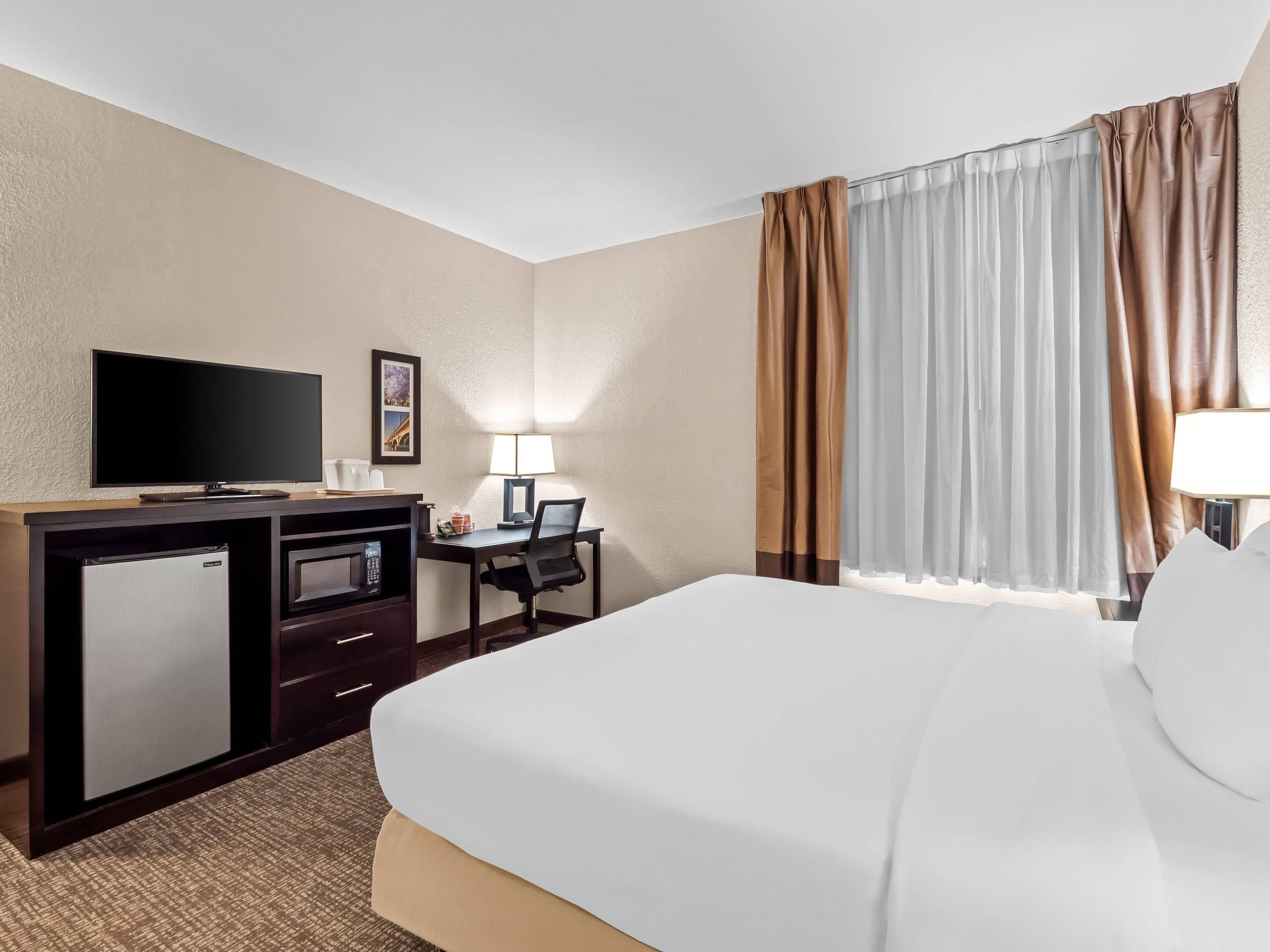 A hotel room with a bed, a desk with a computer, a television, a mini-fridge, and a lamp.