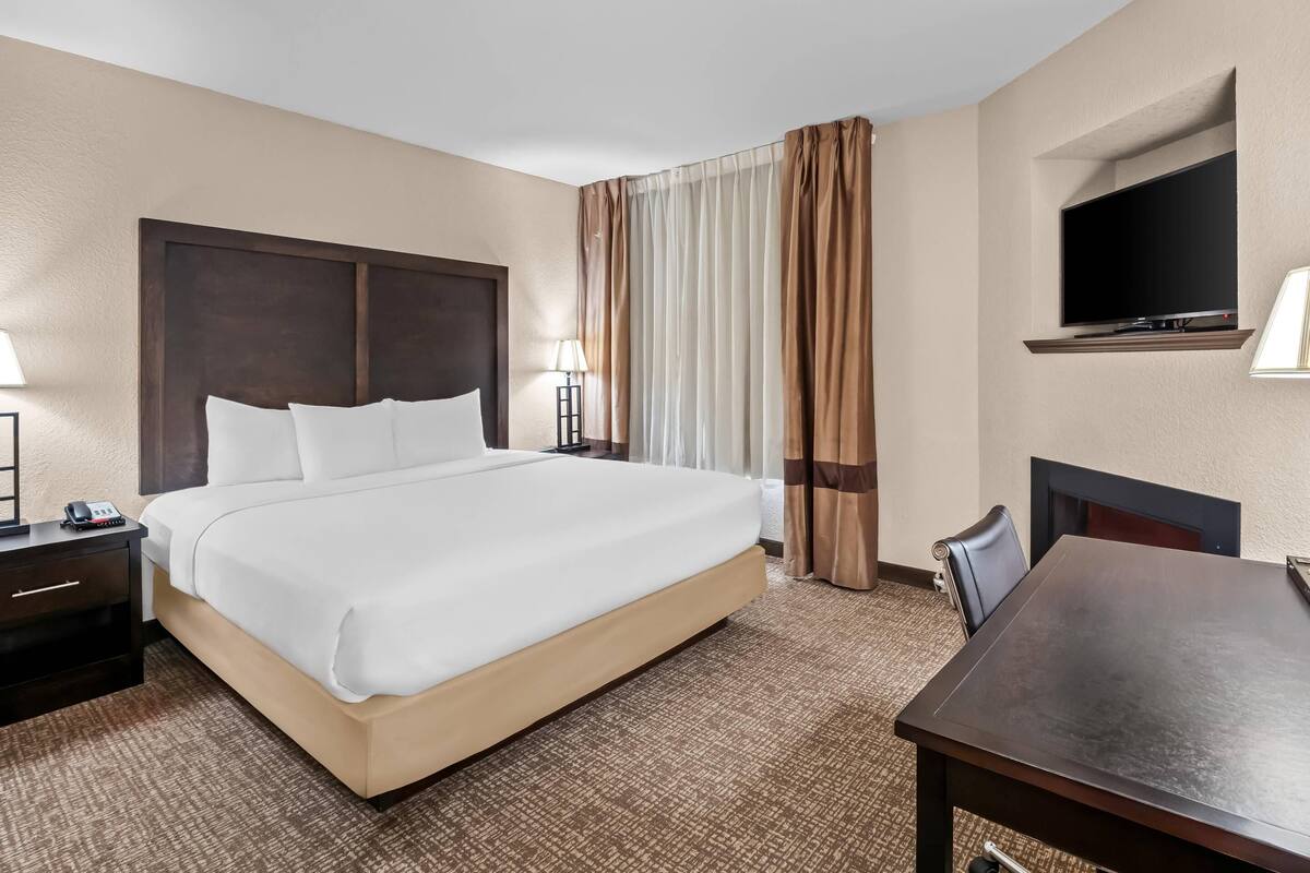 A hotel room with a large bed, a desk, a chair, a TV mounted on the wall, and a window with curtains.