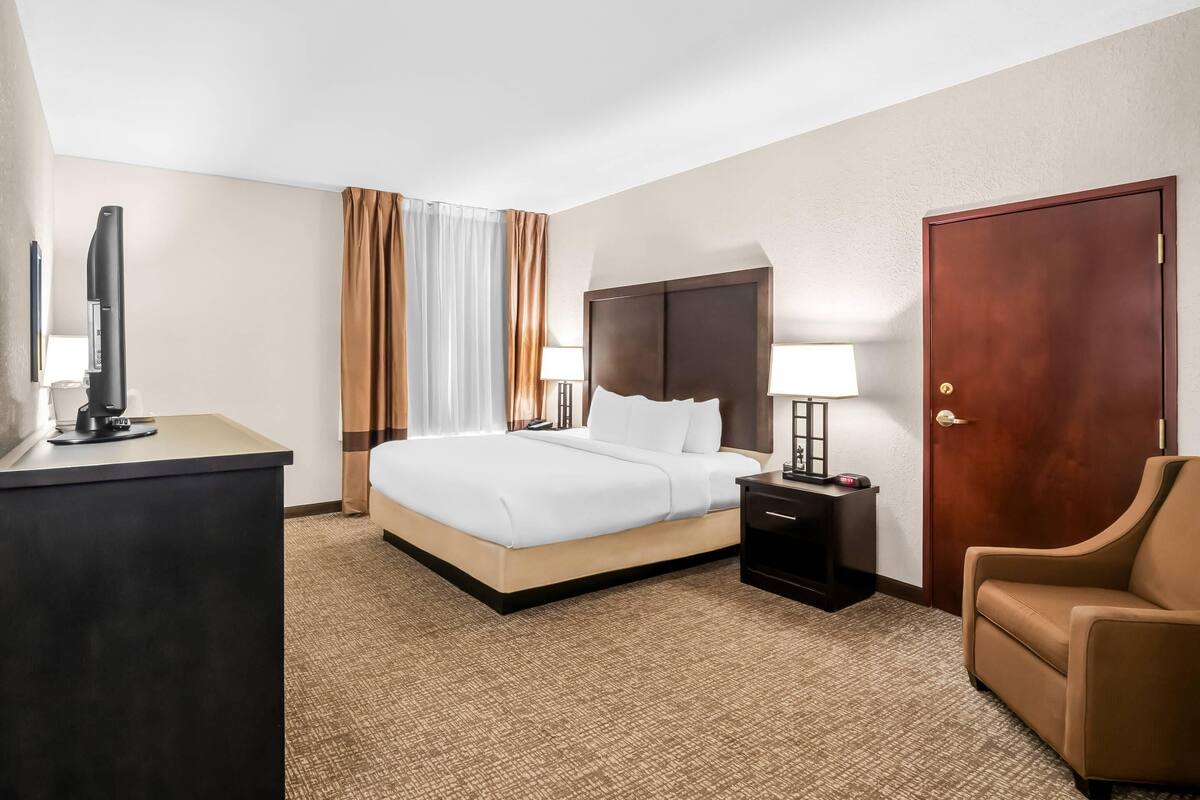 A hotel room with a bed, bedside table, lamp, TV, and a chair.