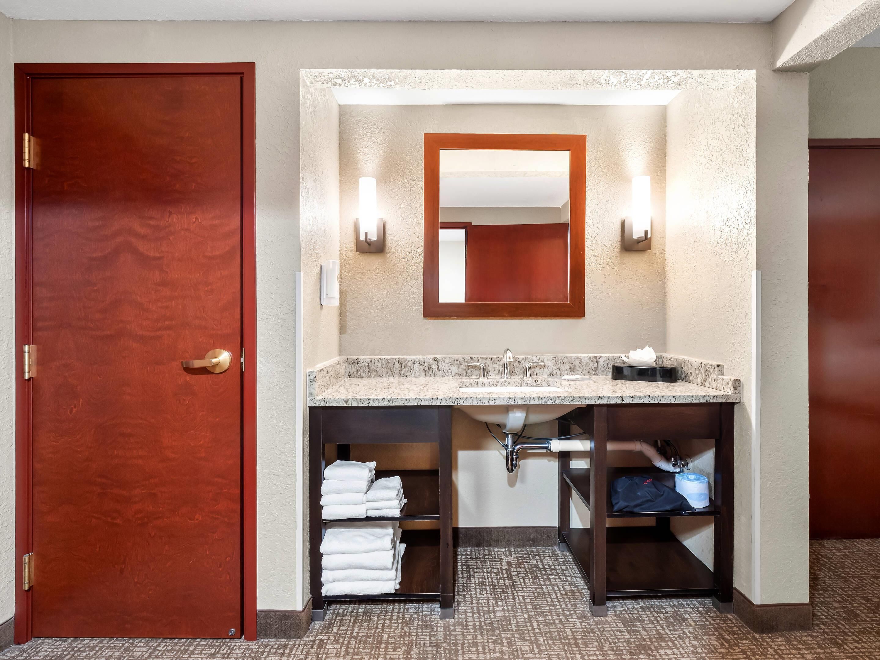 A hotel bathroom with a wooden door, a marble sink, a mirror, and shelves with towels.