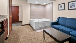 A hotel room with a large bathtub, a blue sofa, a small table, and a kitchenette with a built-in oven. - Comfort Inn & Suites (Allen Park)