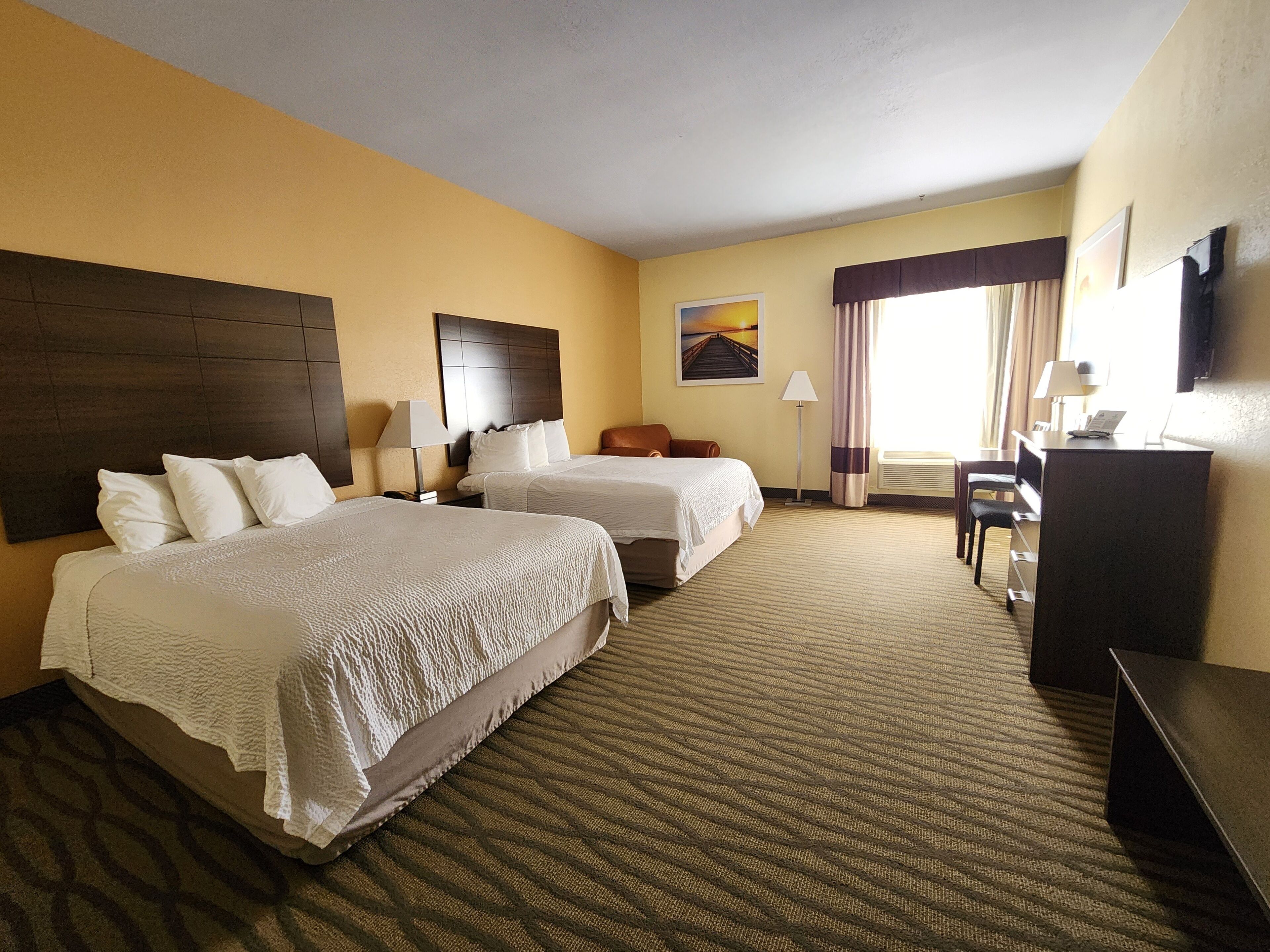 A hotel room with two beds, a desk, a chair, a lamp, and a painting on the wall.
