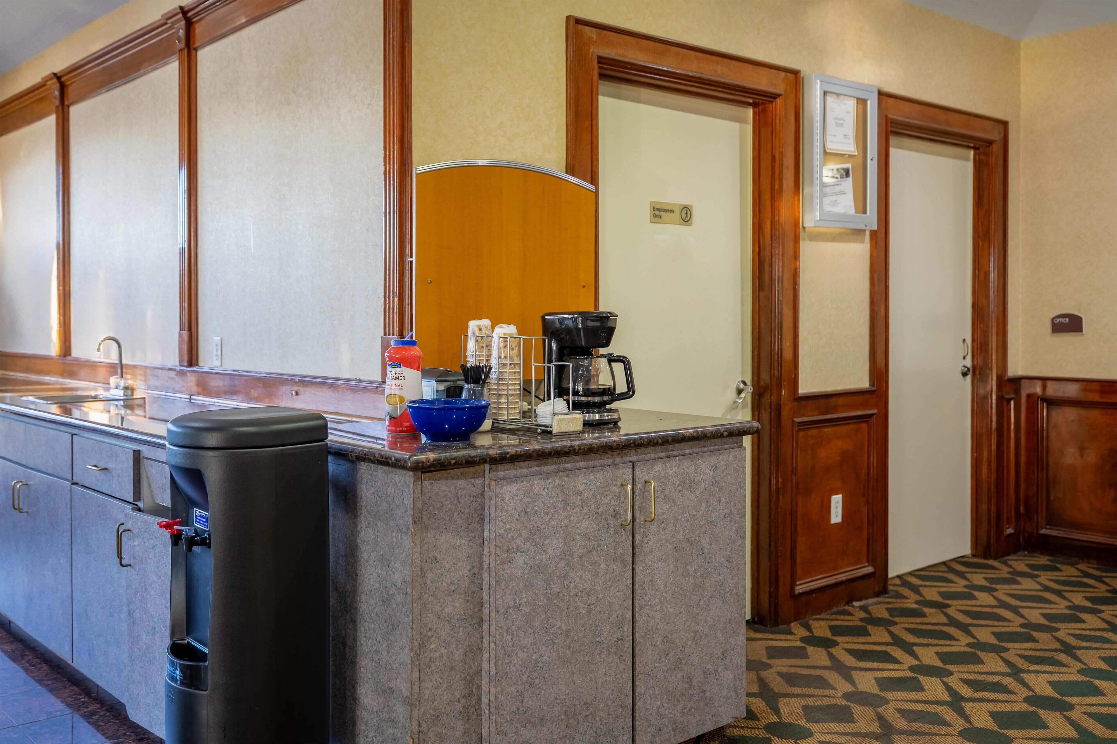Una cocina de hotel con una cafetera, un cubo de basura y un cuenco azul sobre la encimera.