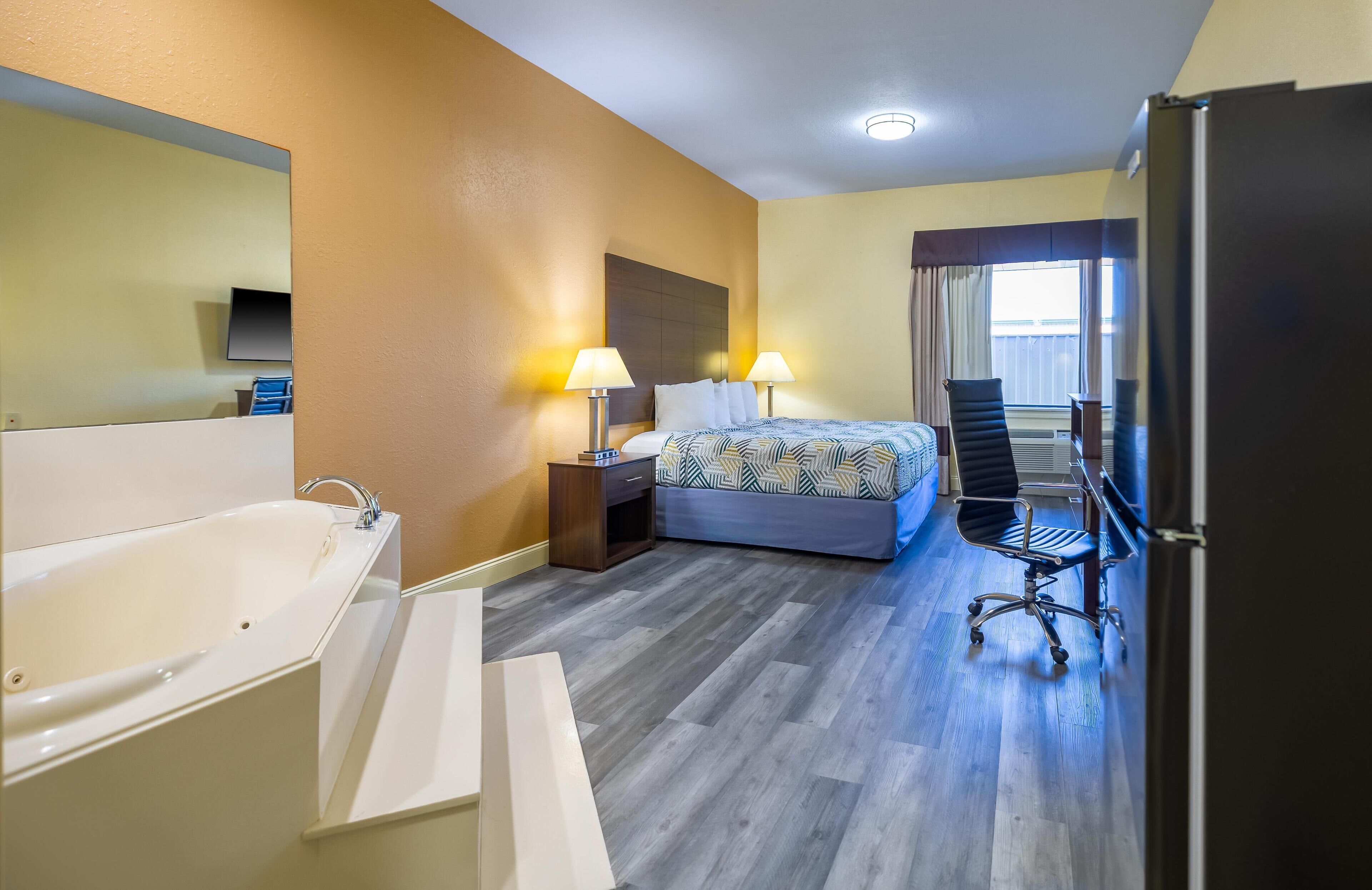 A hotel room with a large bed, a sitting area with a chair, a desk, a flat-screen TV, a bathtub, and a window with curtains.