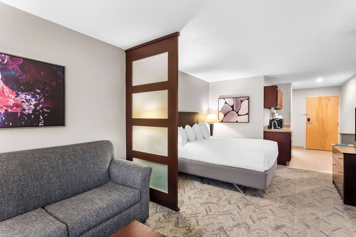A hotel room with a gray sofa, a bed with white bedding, a large abstract painting, a wooden door, and a desk with a lamp.