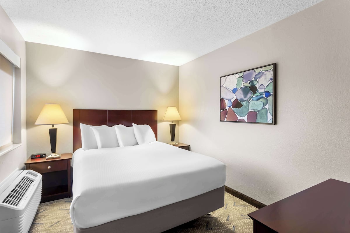 A hotel room with a large bed, two bedside lamps, a digital clock, a heater, and a framed abstract artwork on the wall.