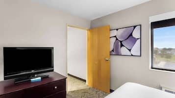 A hotel room with a flat-screen TV on a wooden dresser, a bed, a door leading to another room, and a window with a view.