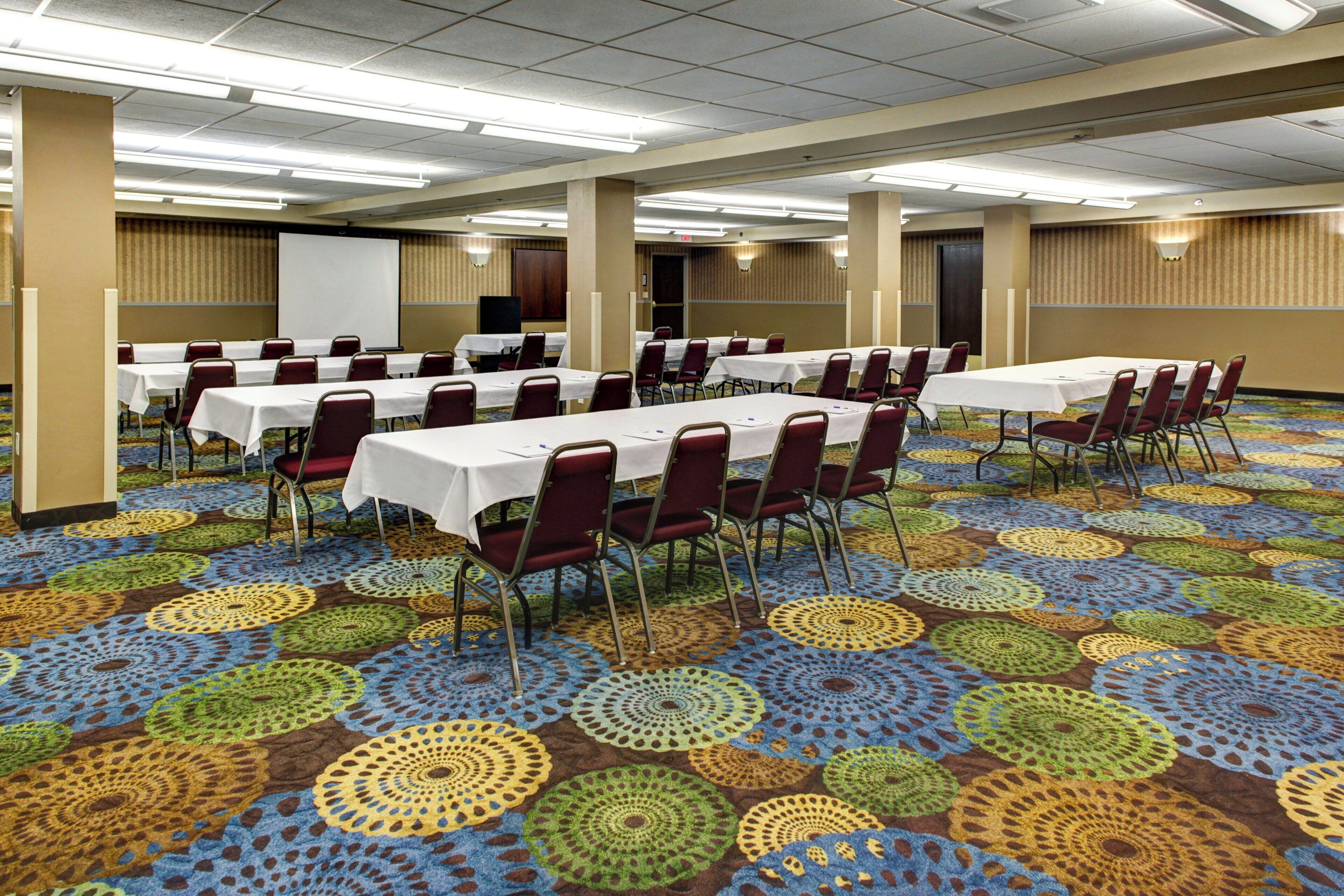 Une salle de conférence équipée de plusieurs tables et chaises, d’un écran de projection et d’un tapis orné d’un motif circulaire.