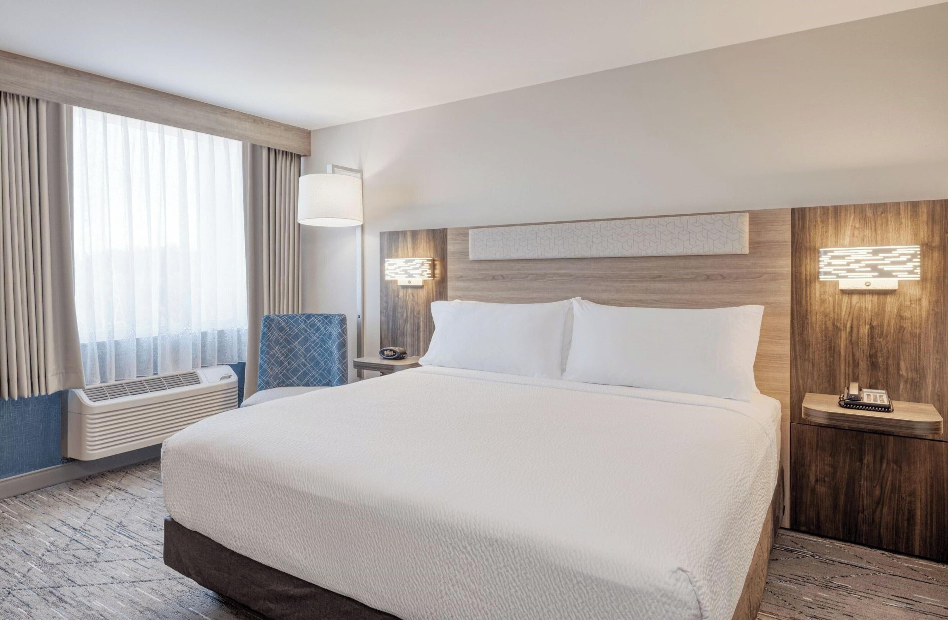 A hotel room with a large bed, a bedside table with a lamp, a wall-mounted light, a window with curtains, and a built-in air conditioning unit.