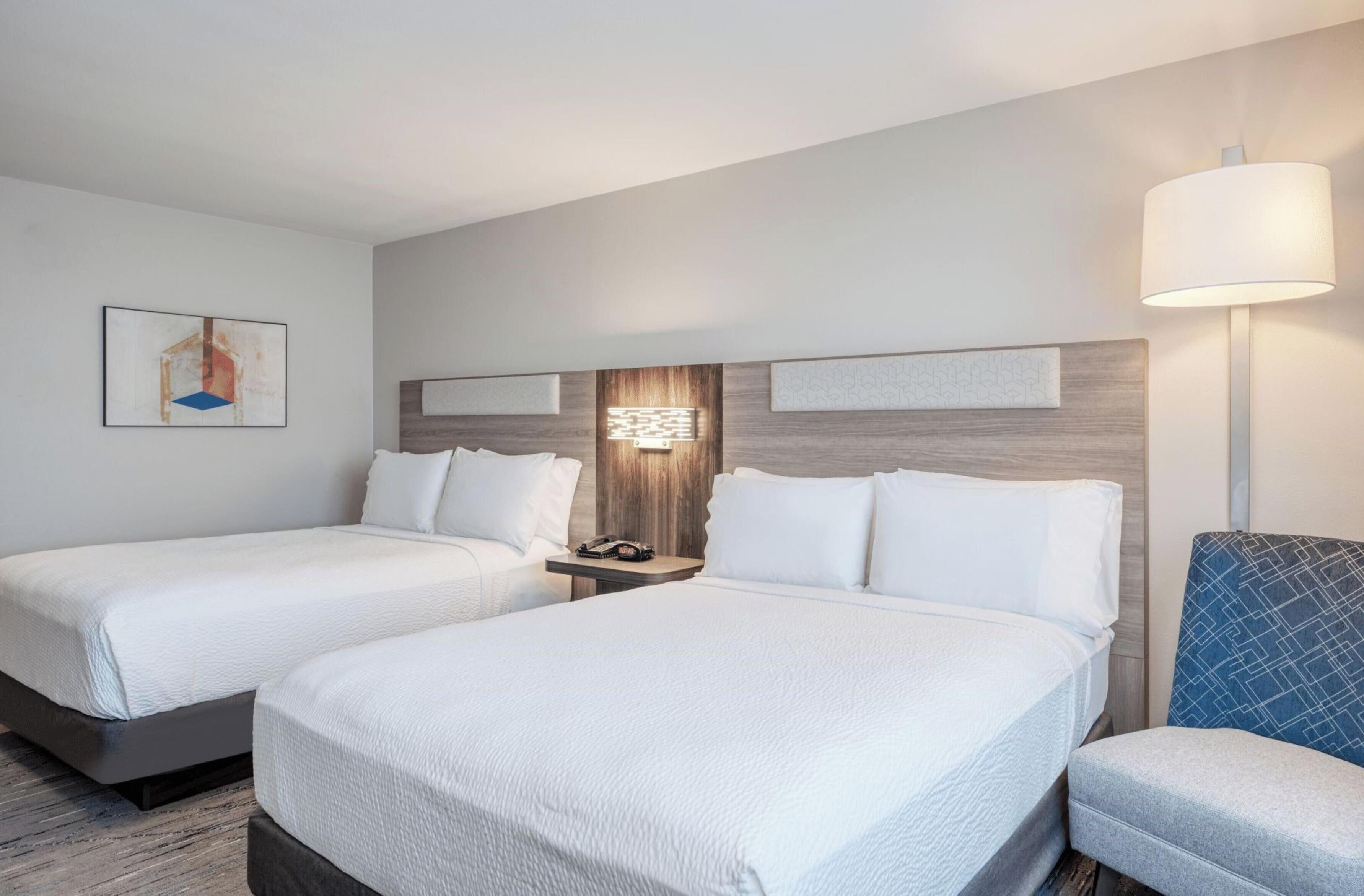 A hotel room with two beds, a nightstand, a lamp, and a chair.