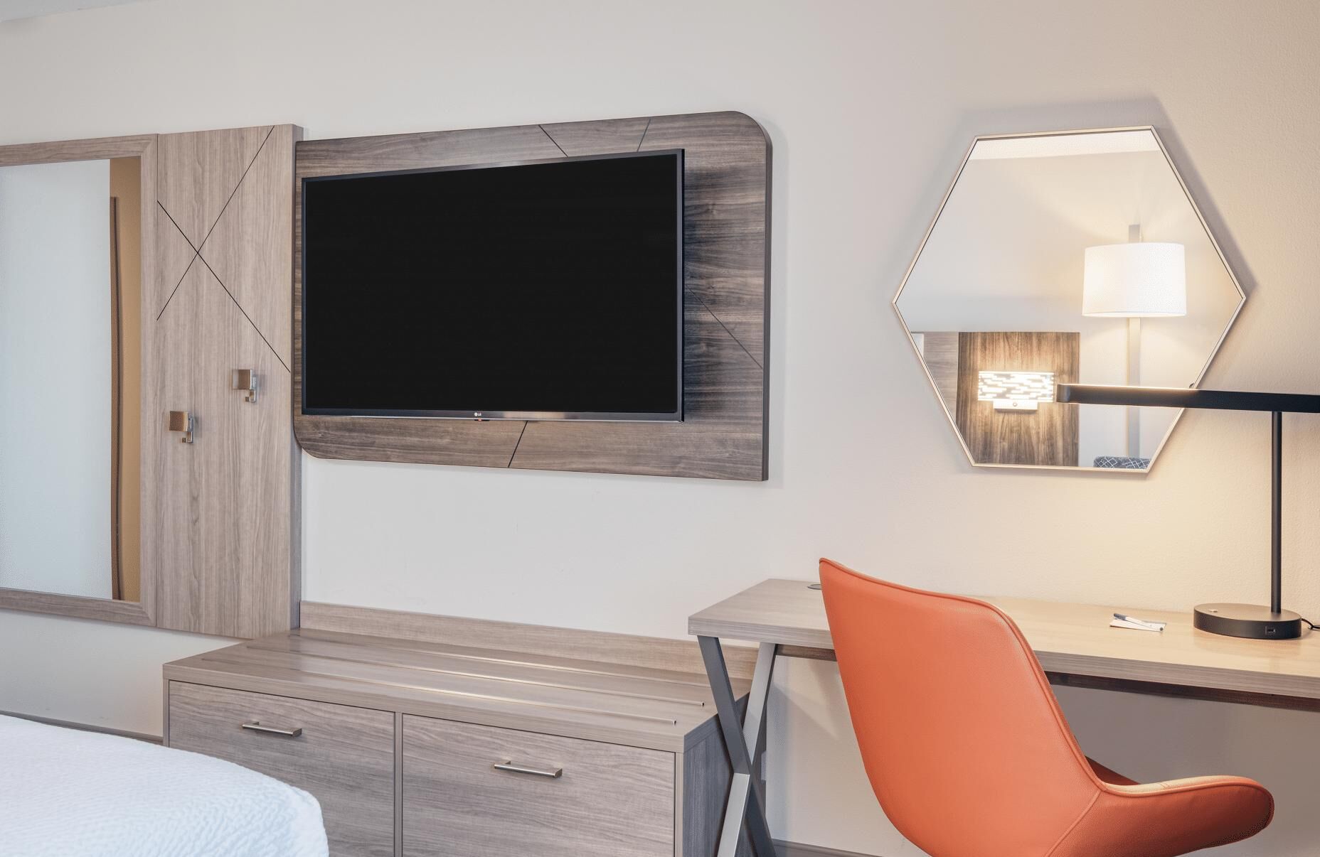 A hotel room with a flat-screen TV mounted on a wooden wall, a desk with a chair, a bedside table, and a mirror with a light.