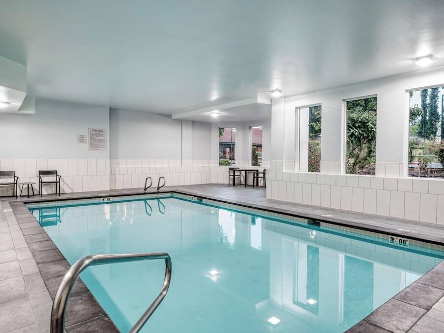 An indoor swimming pool with a capacity sign of 55 users, surrounded by white tiled walls and a ceiling with recessed lighting.