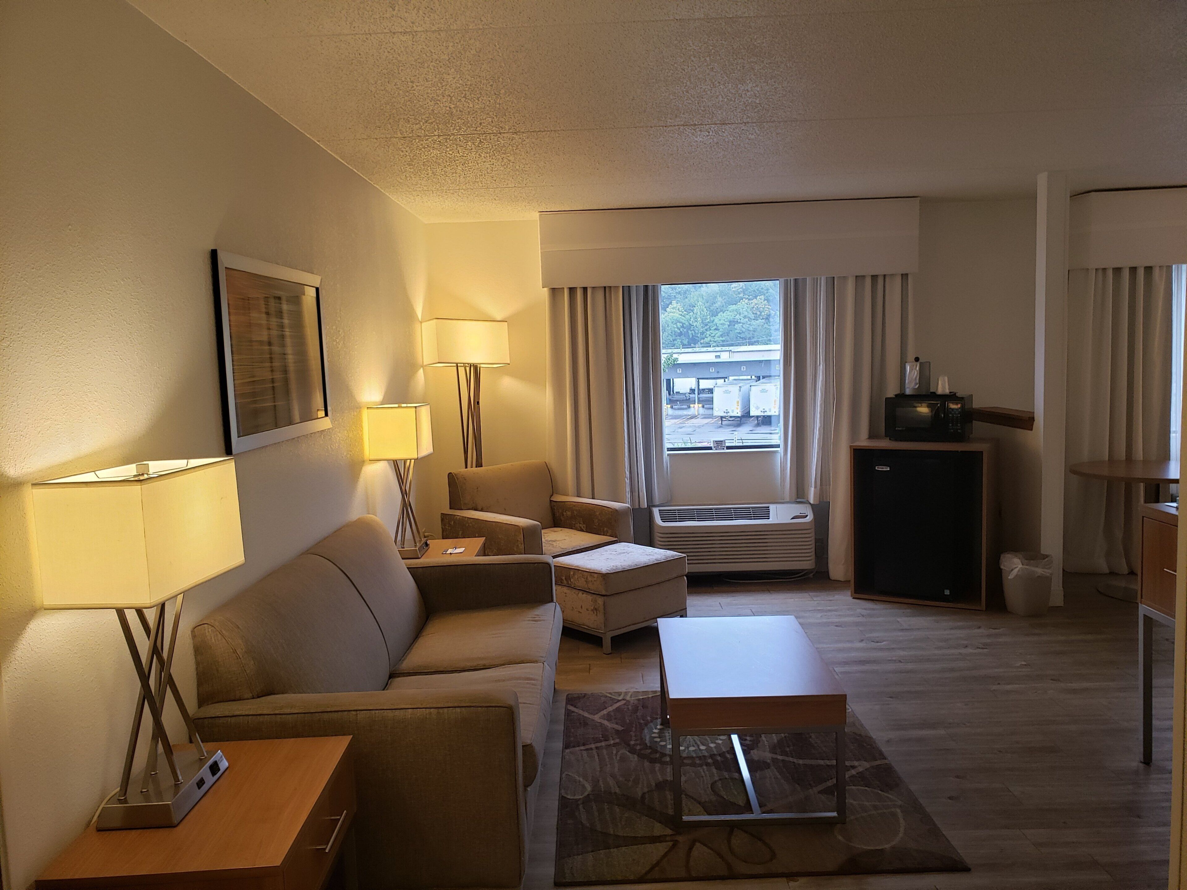 A hotel room with a sofa, armchair, coffee table, side table, lamp, air conditioning unit, and a television on a cabinet.