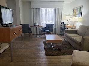 Een hotelkamer met een houten bureau, twee stoelen, een bank, een televisie, een lamp en een raam met gordijnen.