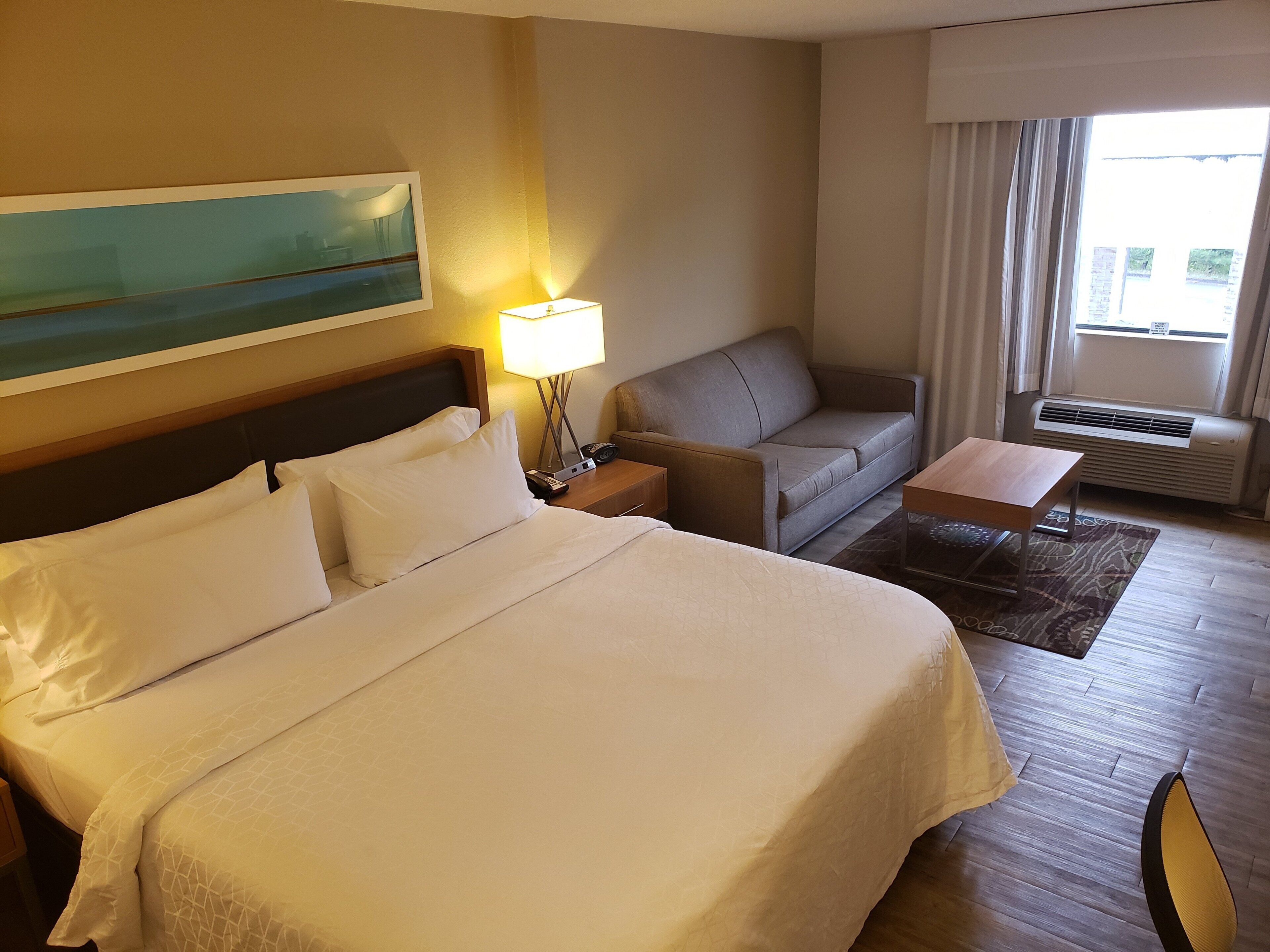 A hotel room with a large bed, a sofa, a small table, a lamp, and a painting on the wall.