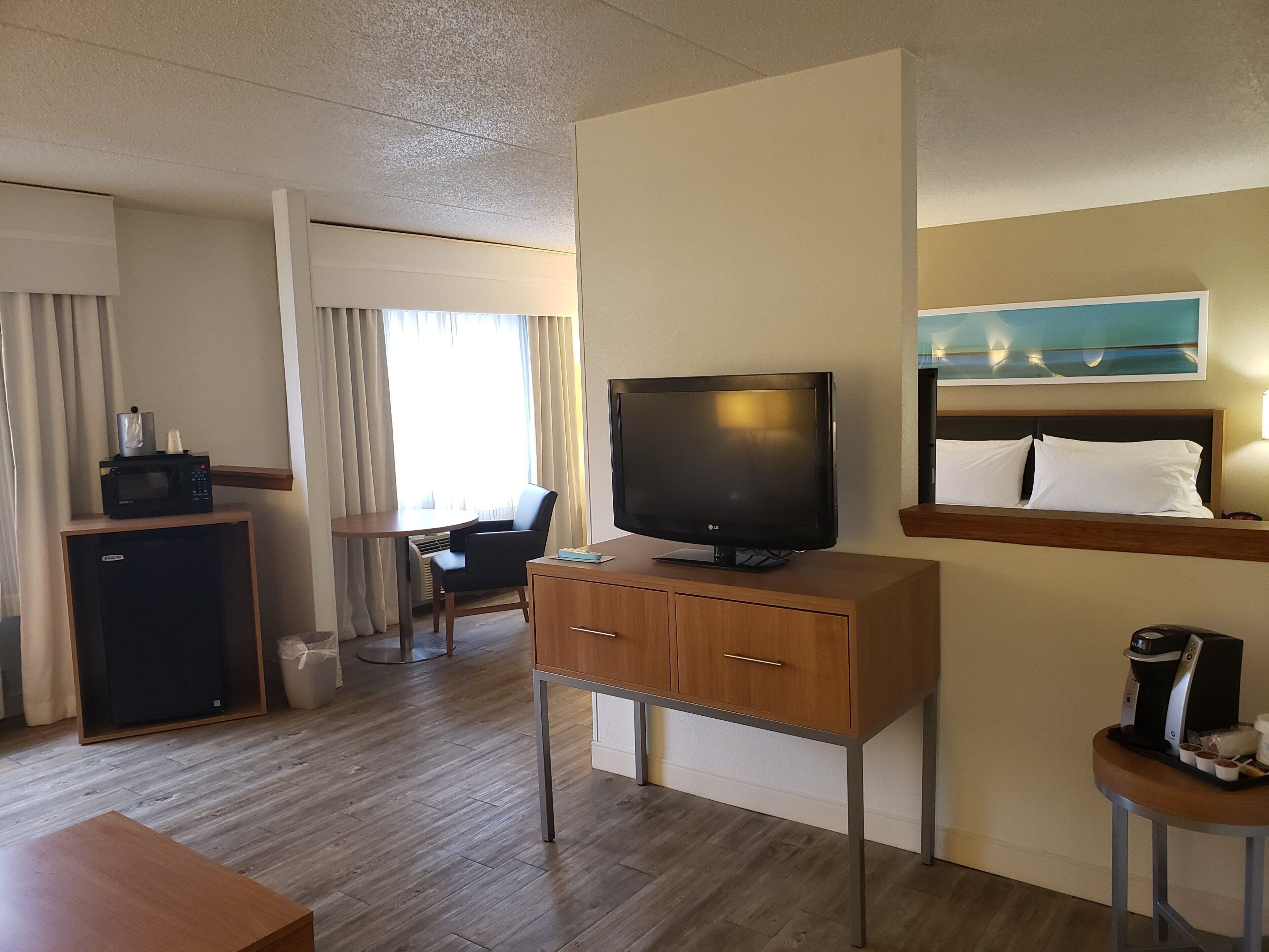 A hotel room with a flat-screen TV, a small desk with drawers, a coffee machine, a bed with white pillows, and a window with curtains.