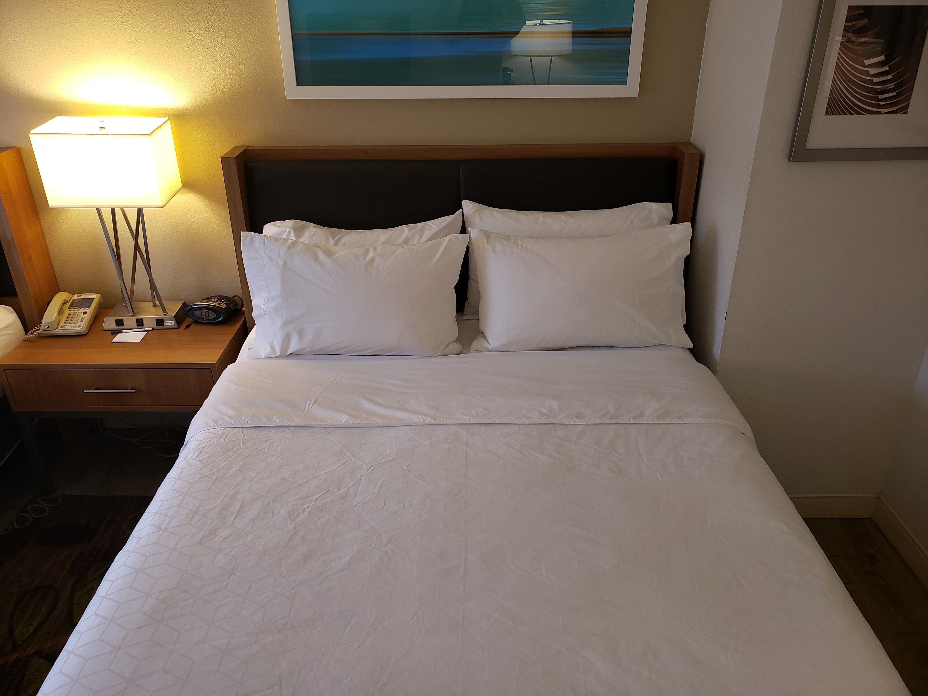 A neatly made bed with white linens, a wooden headboard, a bedside table with a lamp, a telephone, and a framed picture on the wall.