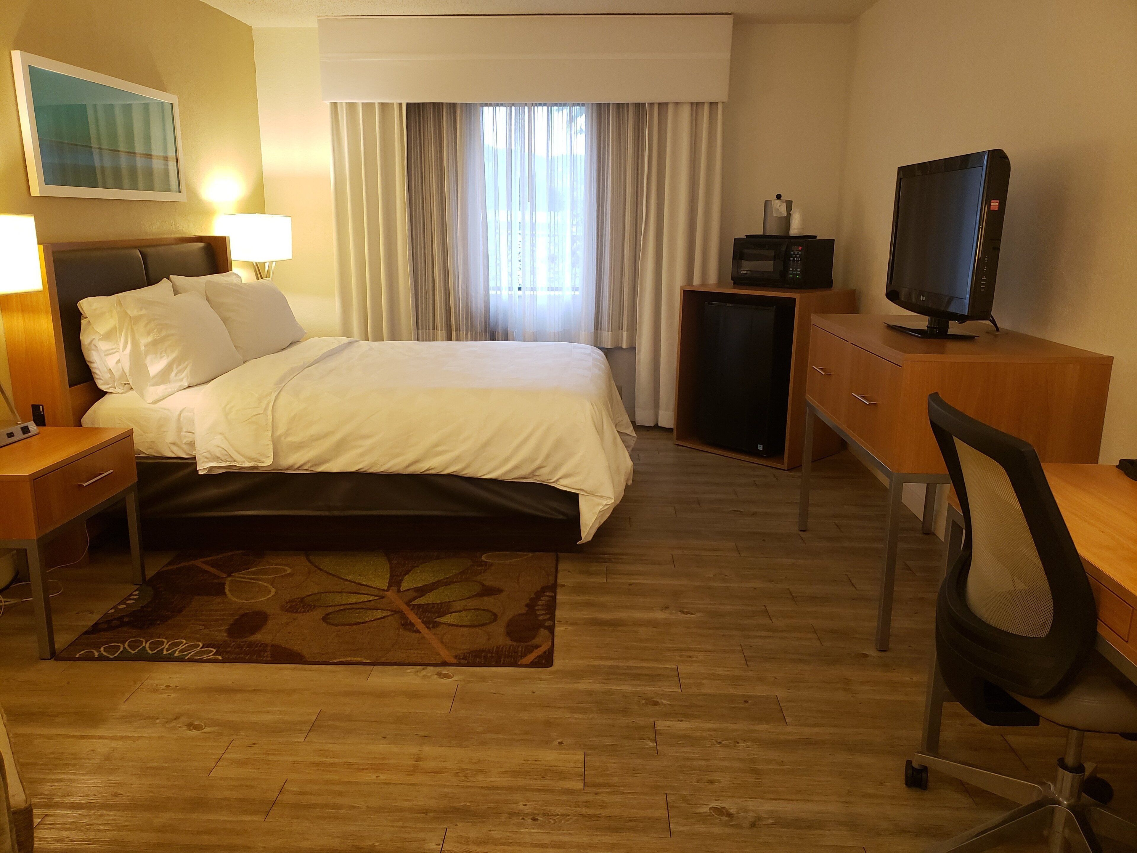 A hotel room with a bed, a desk, a television, a desk lamp, a small cabinet, and a window with curtains.