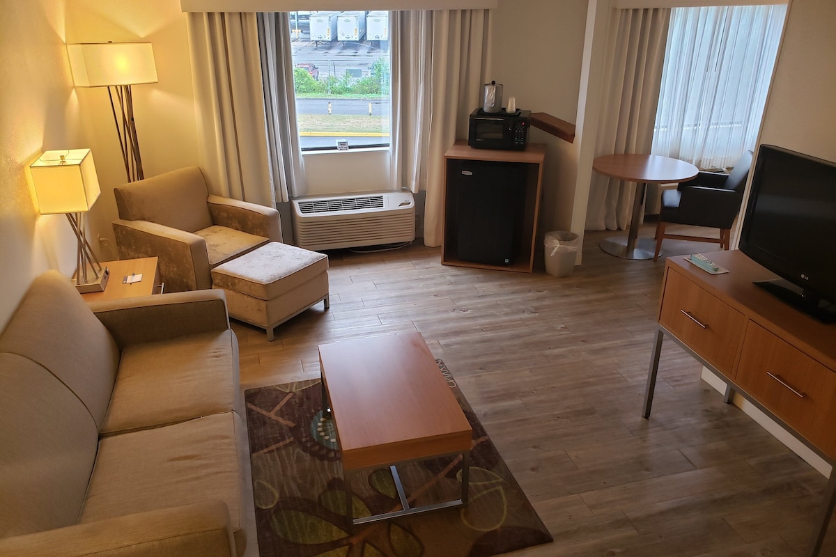 Een hotelkamer met een bank, fauteuil, poef, salontafel, televisie en een klein tafeltje met een lamp.
