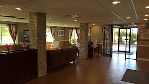 A lobby with stone pillars, a seating area, a reception desk, and a glass door leading outside. - Lux Hotel (Cordele)