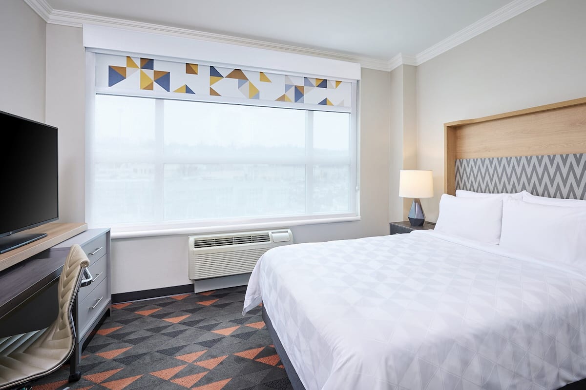 A hotel room with a bed, a TV, a desk, a chair, a lamp, and a window with geometric curtains.