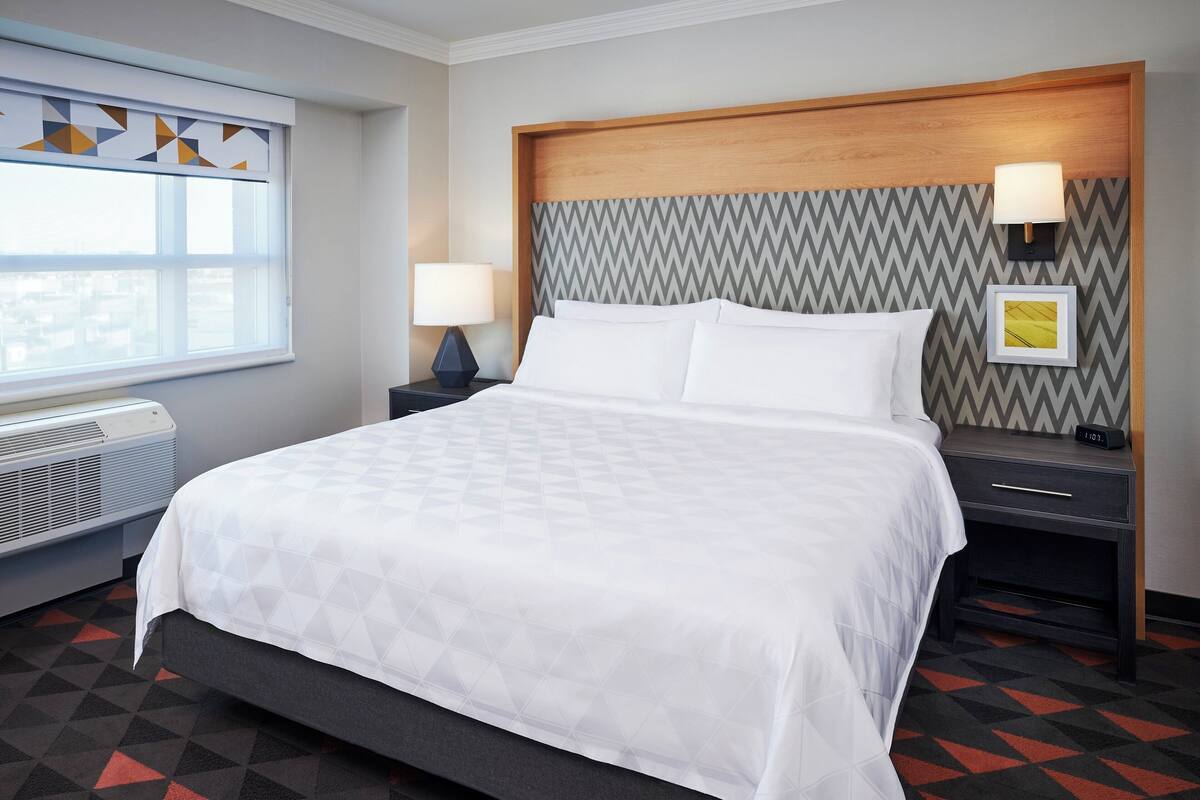 A hotel room with a large bed, a nightstand with a lamp, a wall-mounted picture, and a window with geometric blinds.