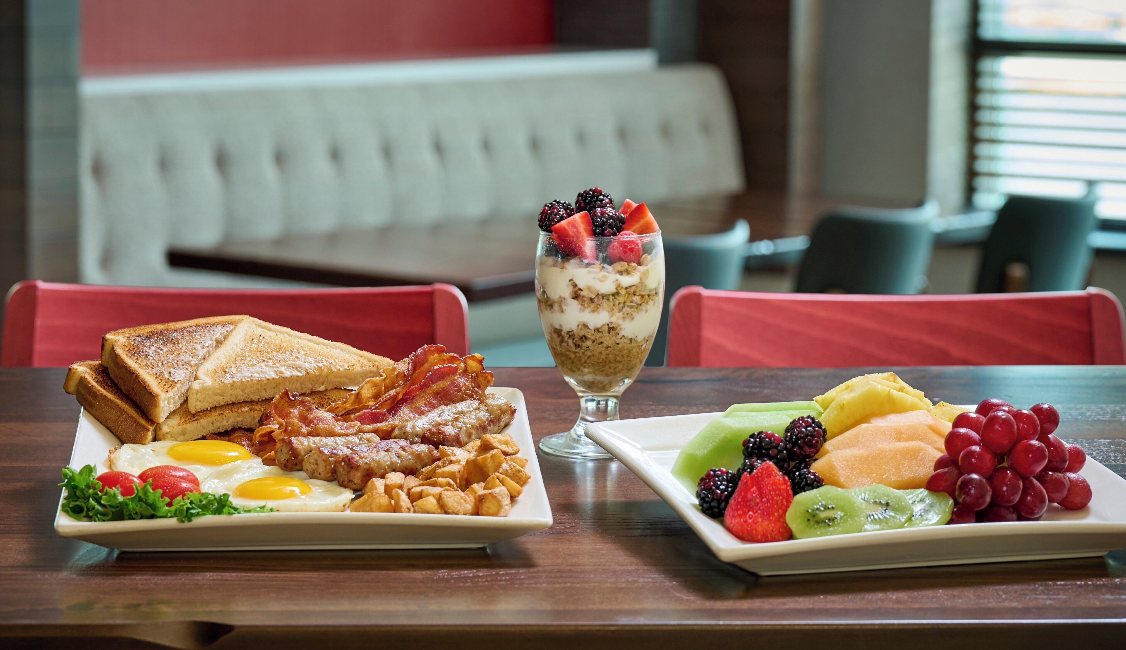 Desayuno con tostadas, huevos, tocino y un parfait de fruta y yogur sobre una mesa.