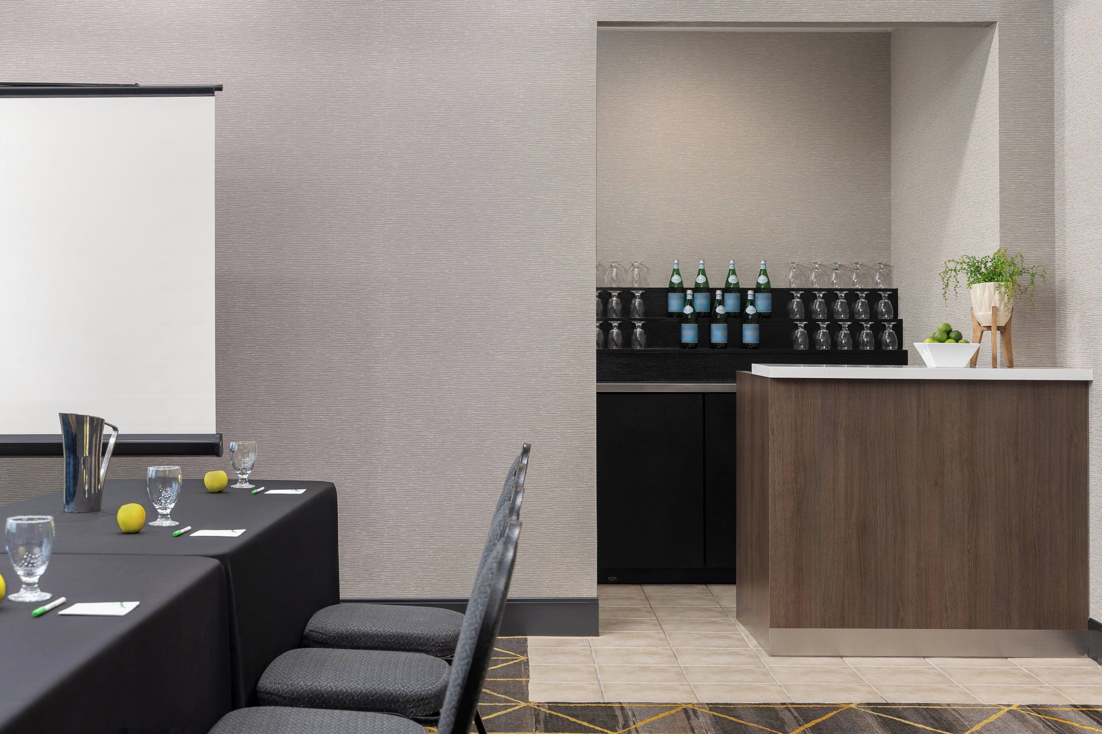 Ein Konferenzraum mit Projektionsleinwand, einem Tisch mit Stühlen, einer kleinen Bar mit Flaschen und Gläsern sowie einem Blumentopf.