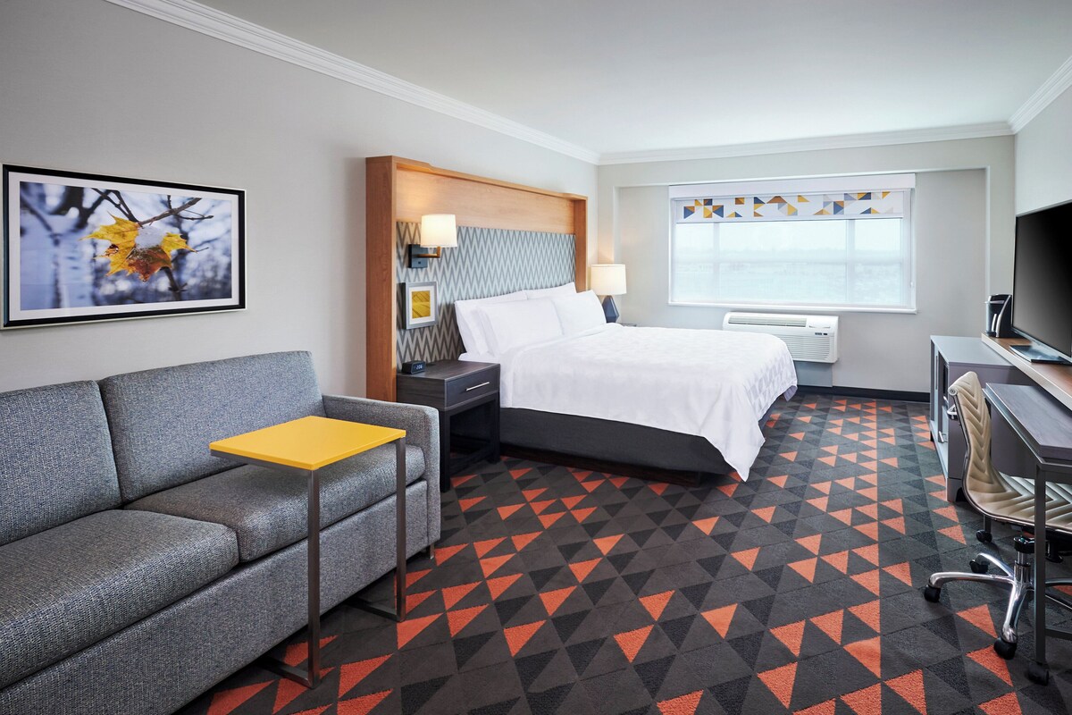 A hotel room with a bed, a desk, a chair, a sofa, a TV, and a framed picture on the wall.