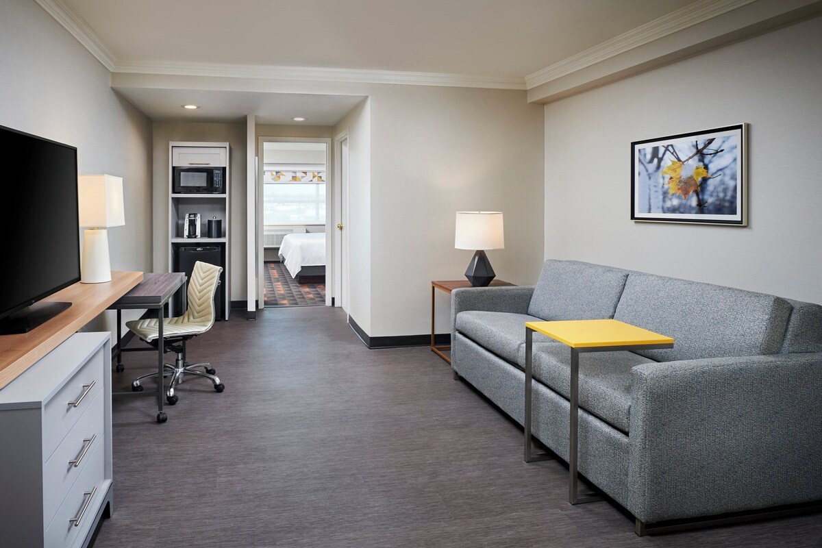 A hotel room with a flat-screen TV, a sofa, a desk with a lamp, a chair, a small table, and a framed picture on the wall.