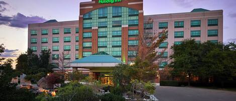 A Holiday Inn hotel with a prominent green logo, a large glass entrance, and a landscaped garden area with yellow flowers.